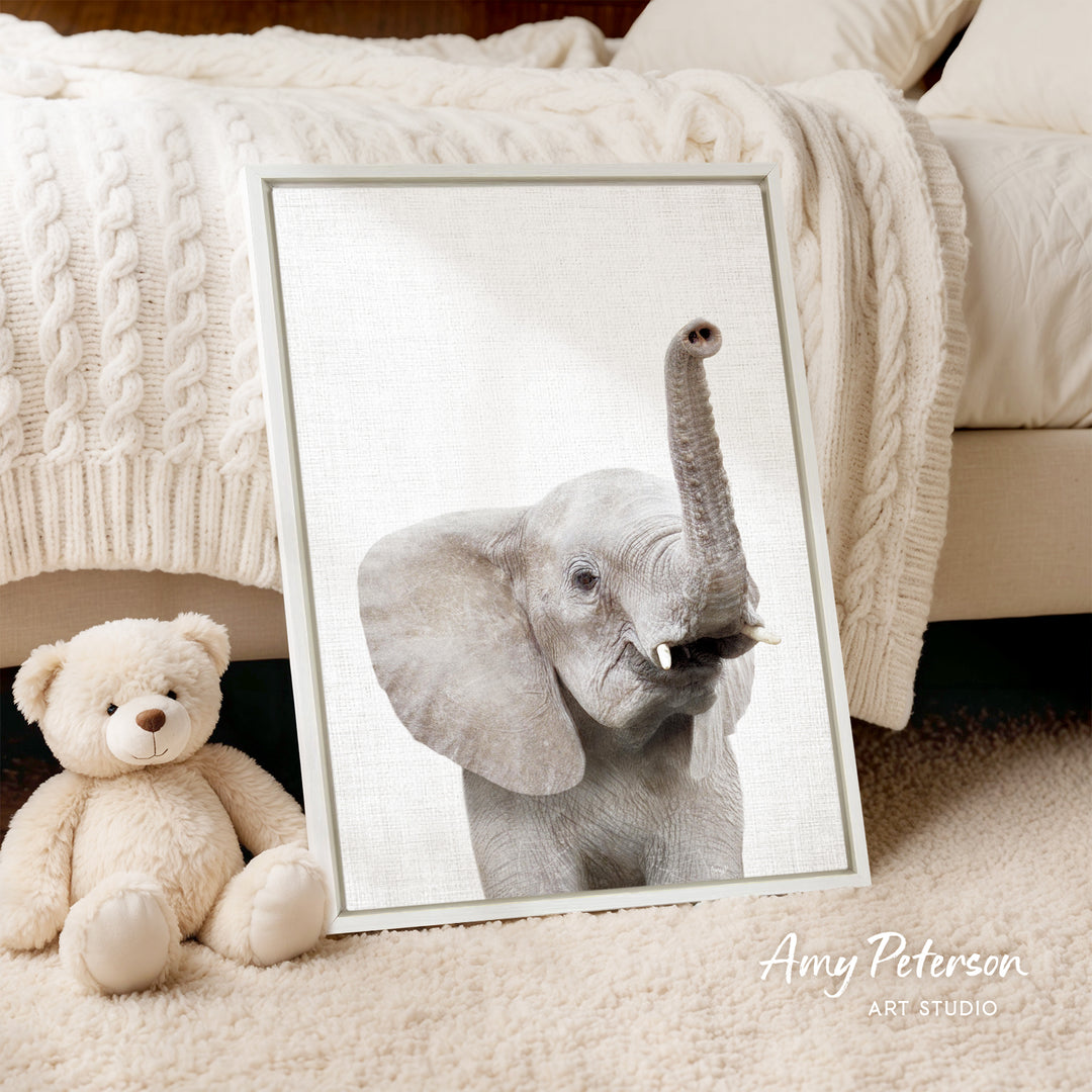A framed photograph of an elephant is displayed next to a teddy bear on a bed, with a white blanket and a wooden headboard in the background.