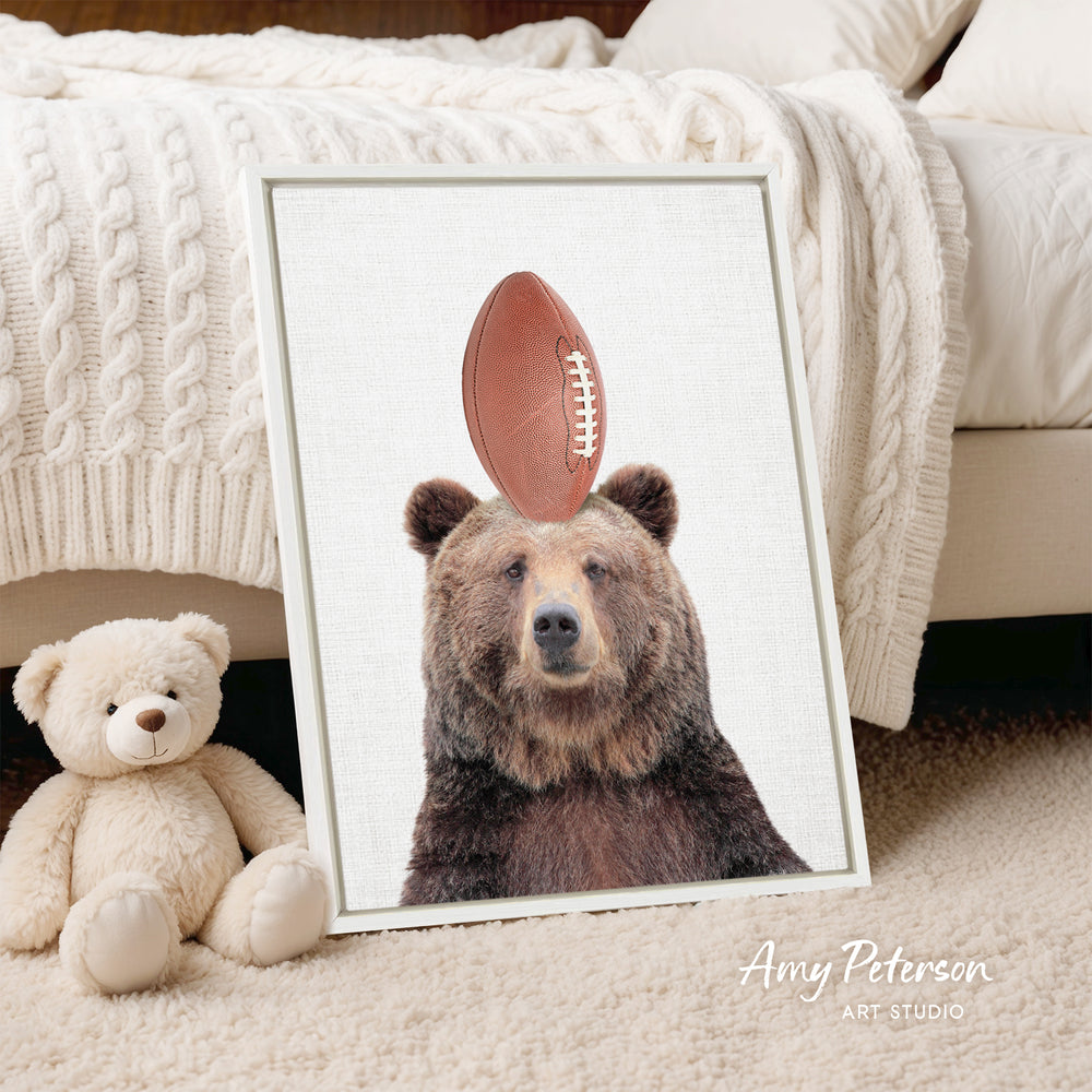 A framed image of a brown bear with a football on its head, placed on a bed next to a stuffed teddy bear.