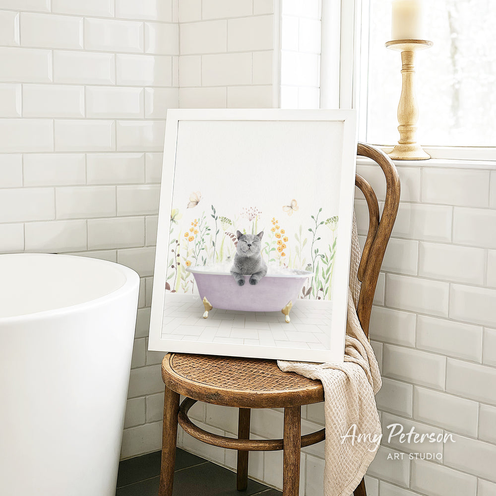 A framed illustration of a cat sitting in a bathtub, surrounded by flowers and butterflies, is displayed on a wooden chair in a bathroom setting.