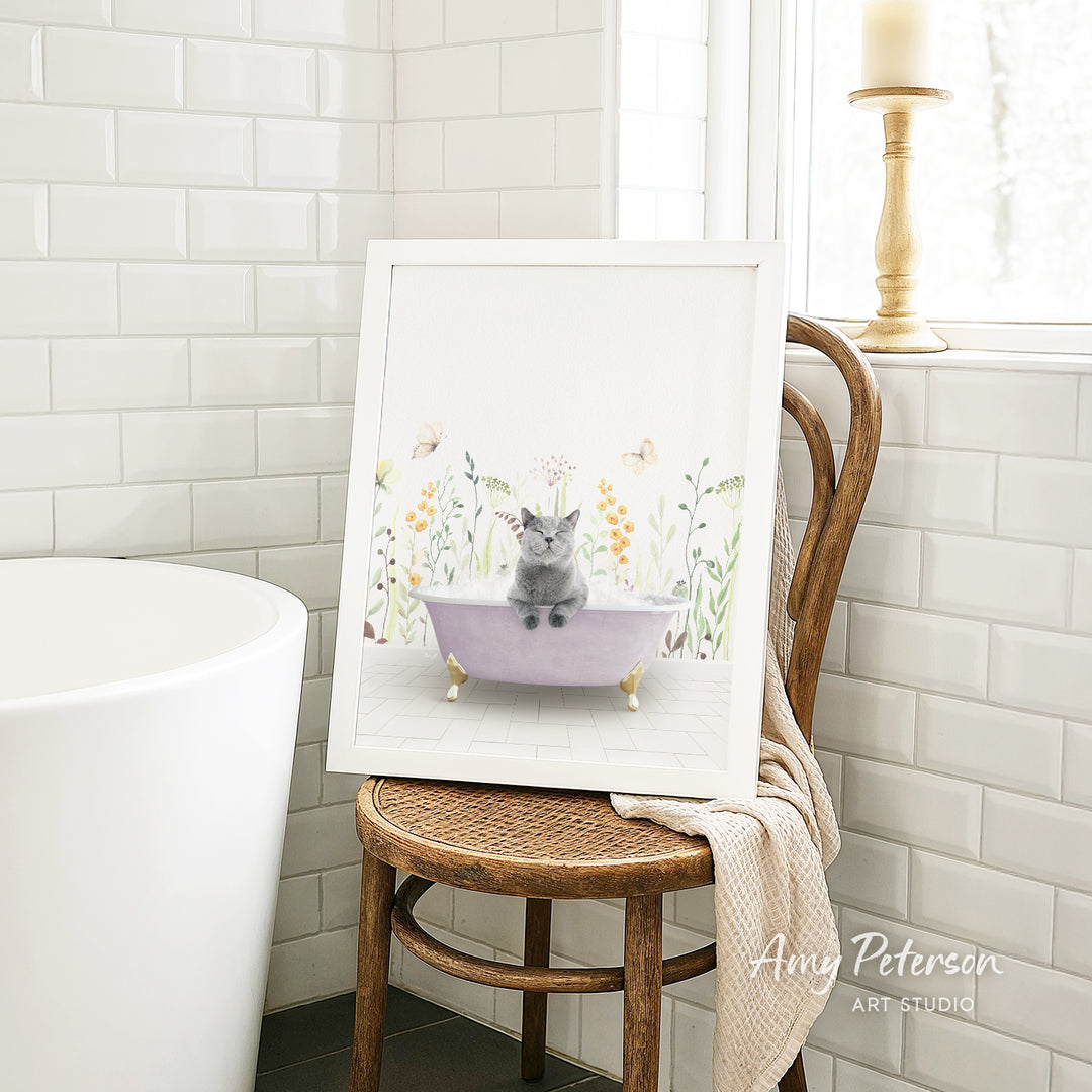 A framed illustration of a cat sitting in a bathtub, surrounded by flowers and butterflies, is displayed on a wooden chair in a bathroom setting.