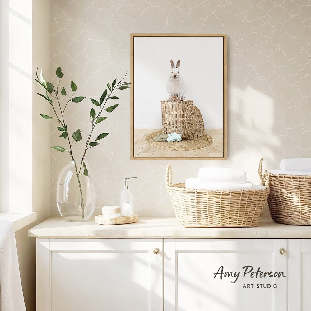 a bathroom scene with a framed picture of a rabbit sitting in a basket, a vase with green leaves, and baskets on a counter.