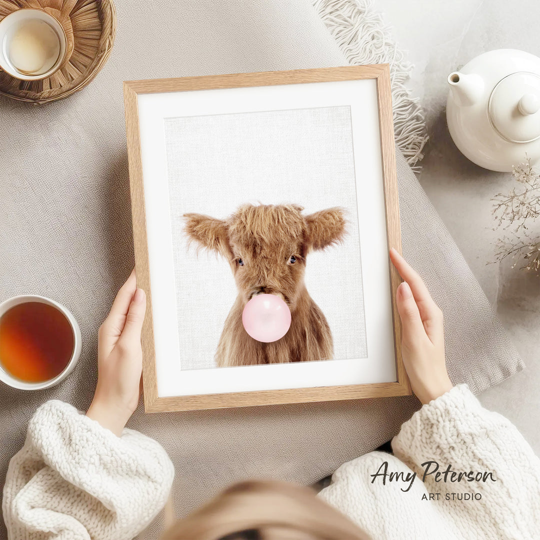 A person is holding a framed picture of a brown cow blowing a bubble with a pink bubble gum bubble.