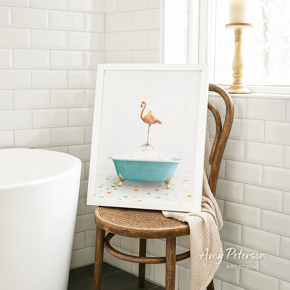 A framed image of a pink flamingo standing in a blue bathtub is displayed on a wooden chair in a bathroom setting.