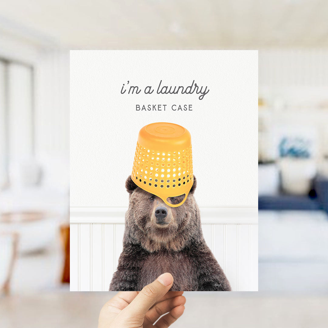A hand holding a picture of a bear wearing a yellow plastic basket on its head, with the caption "I'm a laundry basket case".