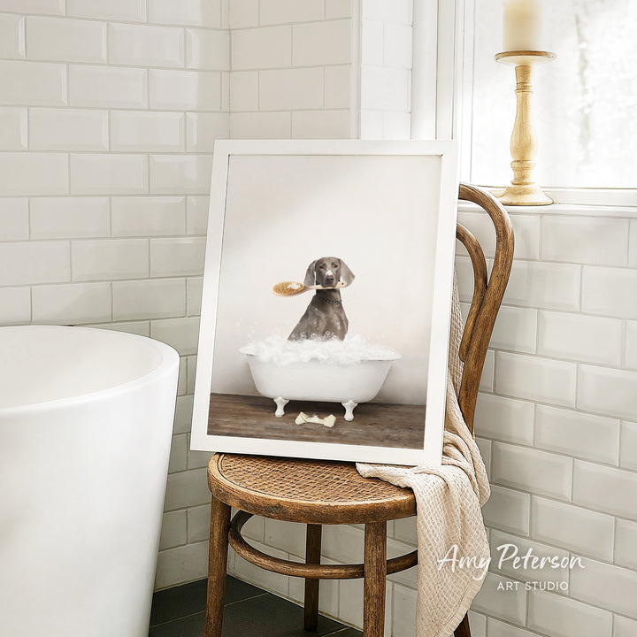 A framed photograph of a dog sitting in a bathtub, with a towel draped over the back of a wooden chair in a bathroom setting.