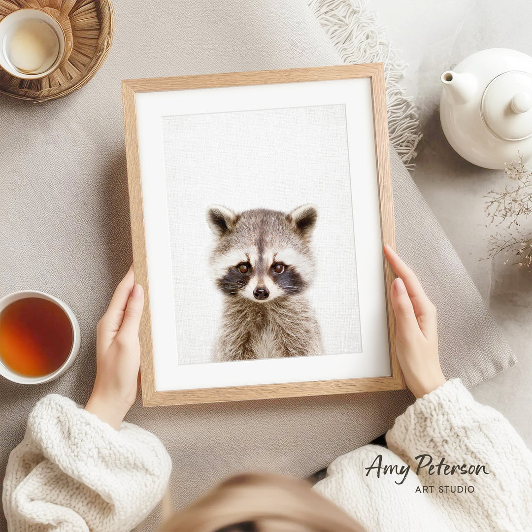 A person is holding a framed picture of a raccoon, with a teapot, a cup of tea, and a bowl of tea visible in the background.