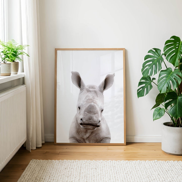 a large framed picture of a rhinoceros, which is the central focus of the room. The rhinoceros is positioned in the center of the frame, and the picture is hanging on a white wall. The room has a wooden floor and is decorated with potted plants and a rug.
