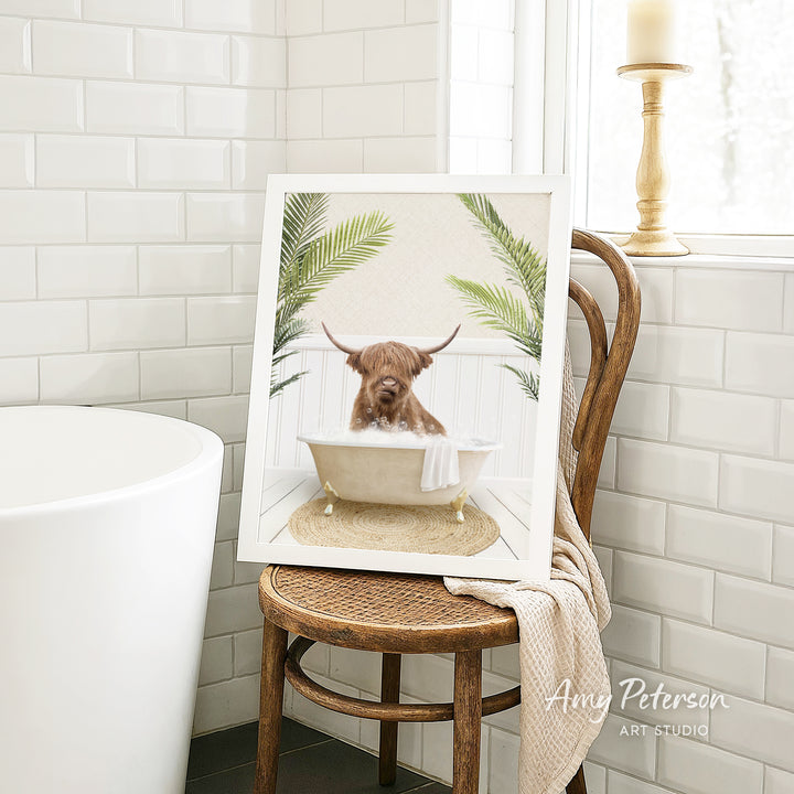 A framed image of a Highland cow sitting in a bathtub, surrounded by tropical plants, is displayed on a wooden chair in a bathroom setting.