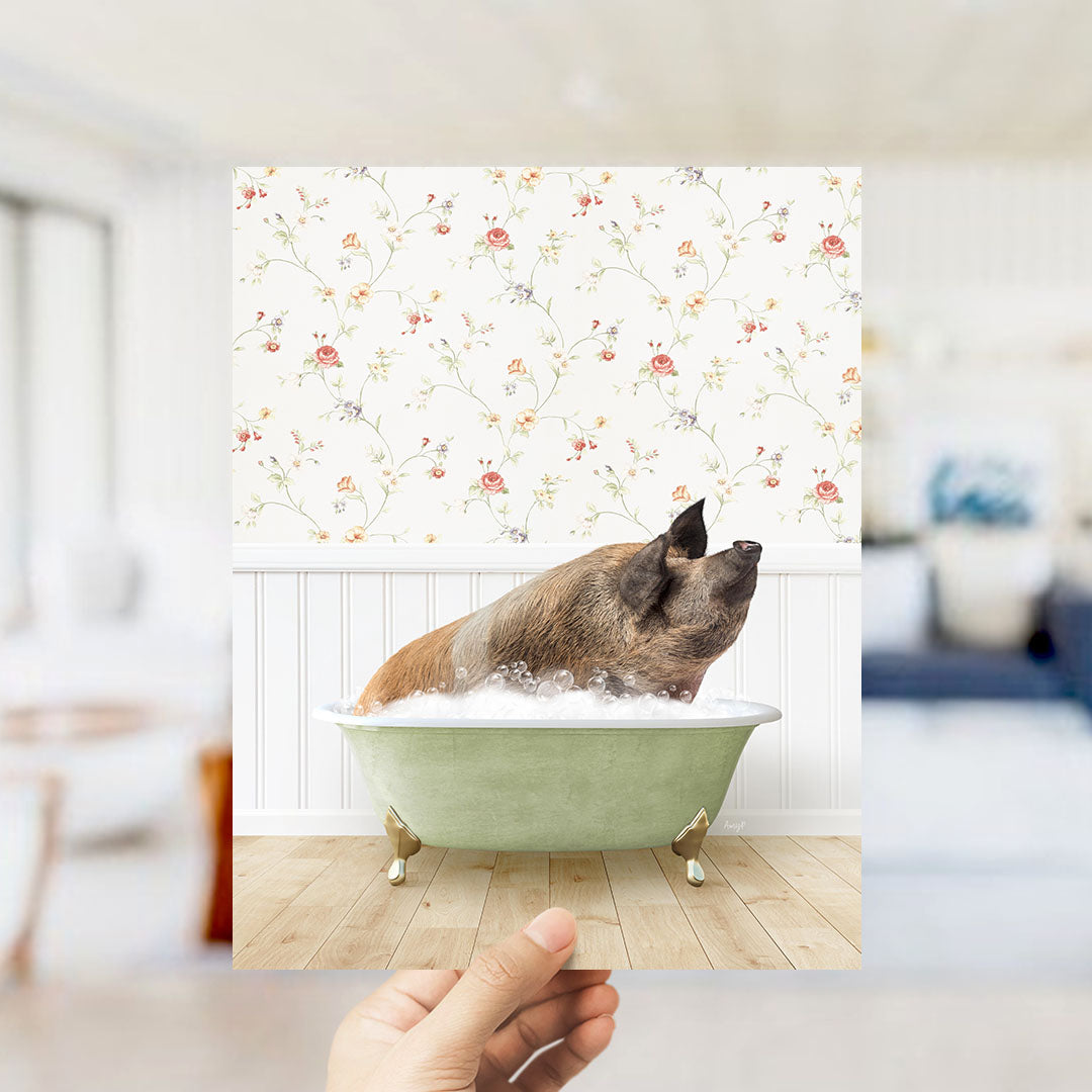 A hand holds up a framed photograph of a pig sitting in a green bathtub filled with bubbles, against a floral wallpaper backdrop.