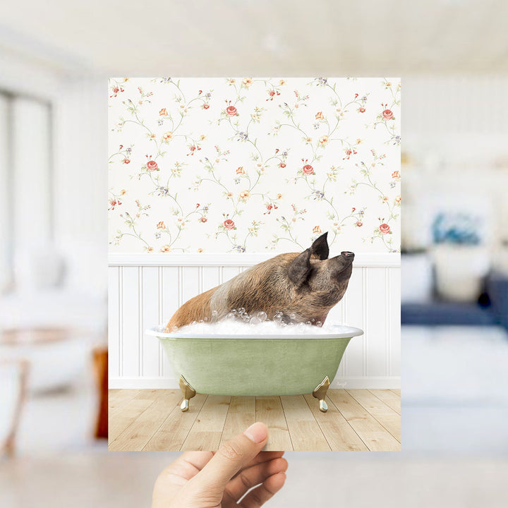 A hand holds up a framed photograph of a pig sitting in a green bathtub filled with bubbles, against a floral wallpaper backdrop.