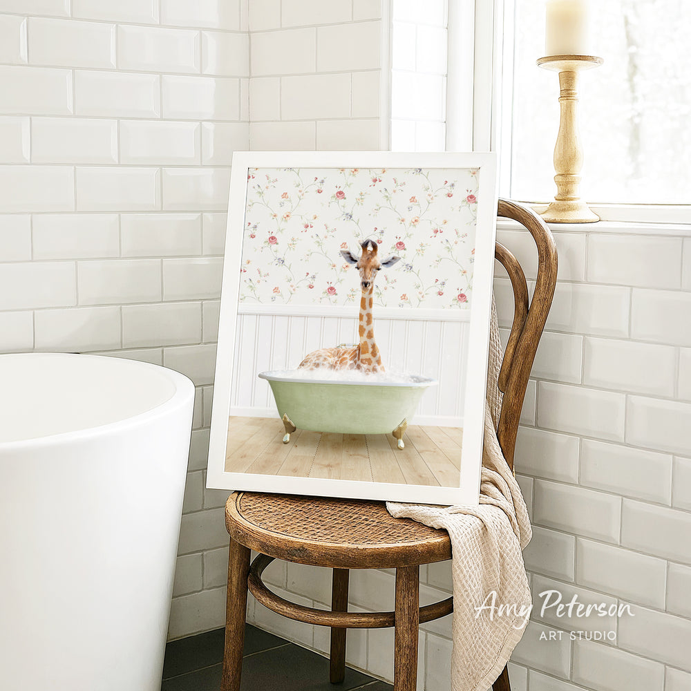 A framed image of a giraffe sitting in a bathtub, surrounded by a floral pattern, is displayed on a wooden chair in a bathroom setting.