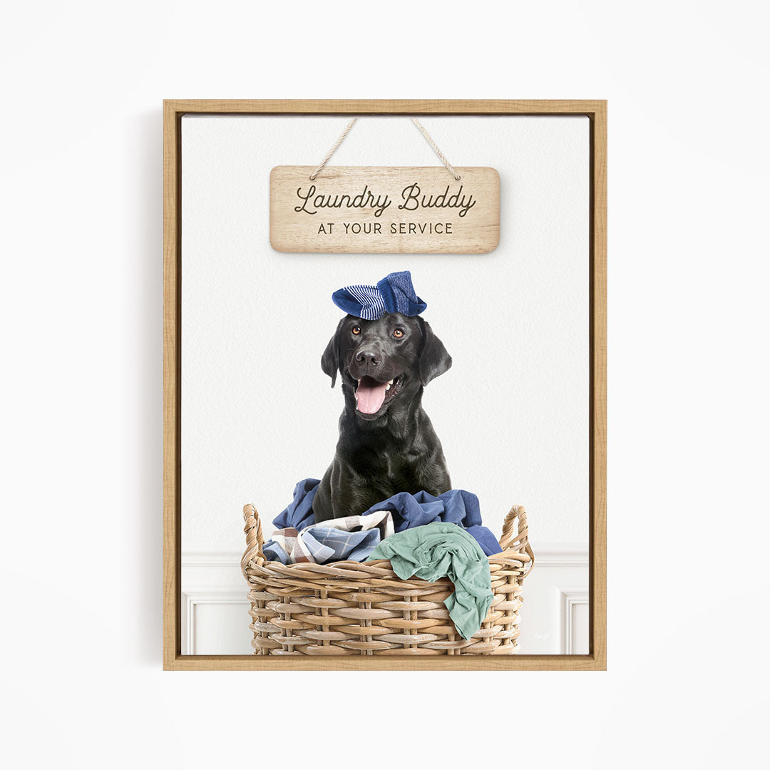 A black dog wearing a blue hat sits in a wicker basket filled with clothes, with a wooden sign above that reads "Laundry Buddy at your service".