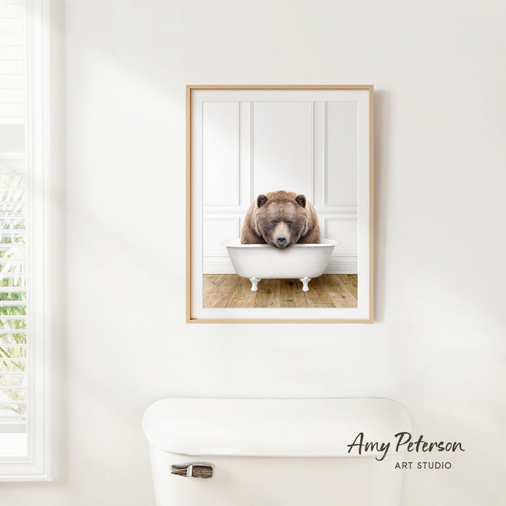 A framed image of a brown bear sitting in a white bathtub, with a window and toilet visible in the background.