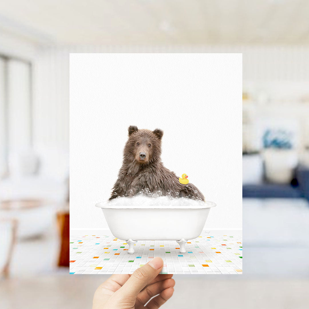 A hand holds up a framed image of a brown bear sitting in a bathtub filled with bubbles, with a rubber duck floating nearby.