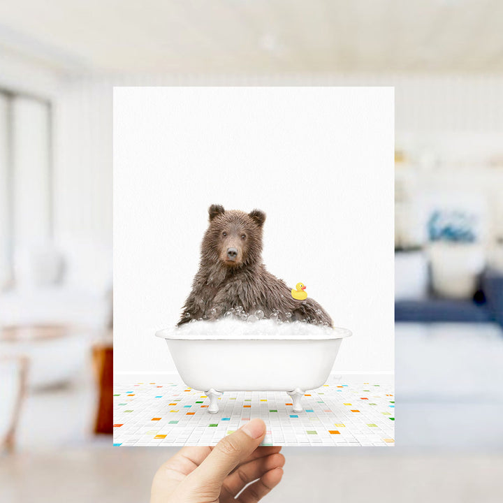A hand holds up a framed image of a brown bear sitting in a bathtub filled with bubbles, with a rubber duck floating nearby.