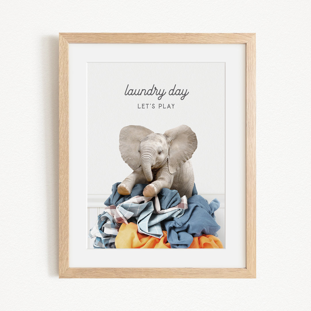 A framed image of a stuffed elephant sitting atop a pile of laundry, with the text "laundry day let's play" written above it.