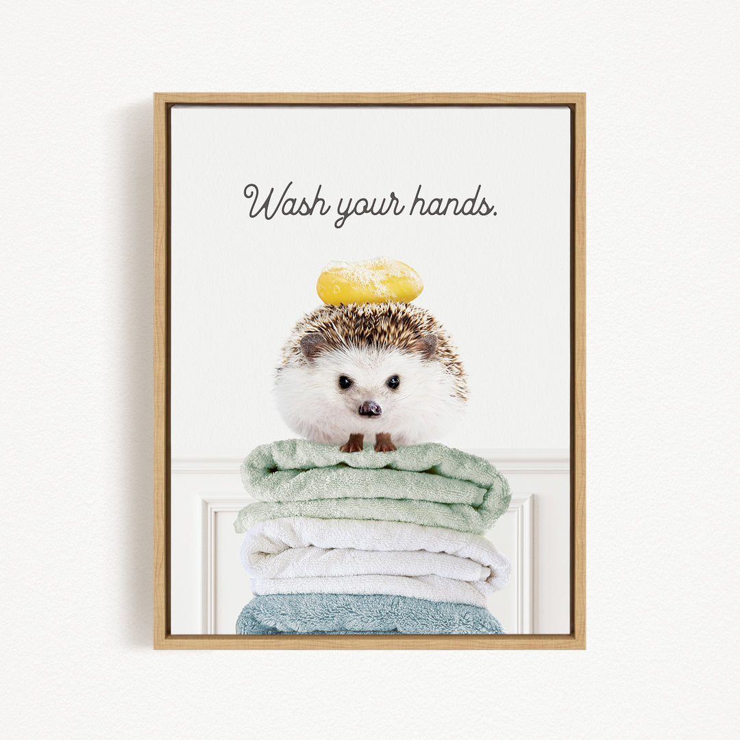 A hedgehog sitting on a stack of folded towels with a yellow object on its head, and the text "Wash your hands" written above it.