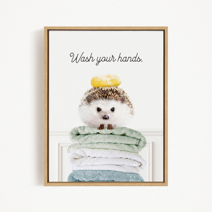 A hedgehog sitting on a stack of folded towels with a yellow object on its head, and the text "Wash your hands" written above it.