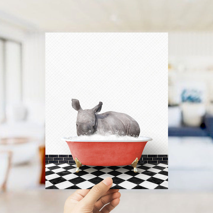 A hand is holding up a framed image depicting a rhinoceros sitting in a red bathtub filled with bubbles.