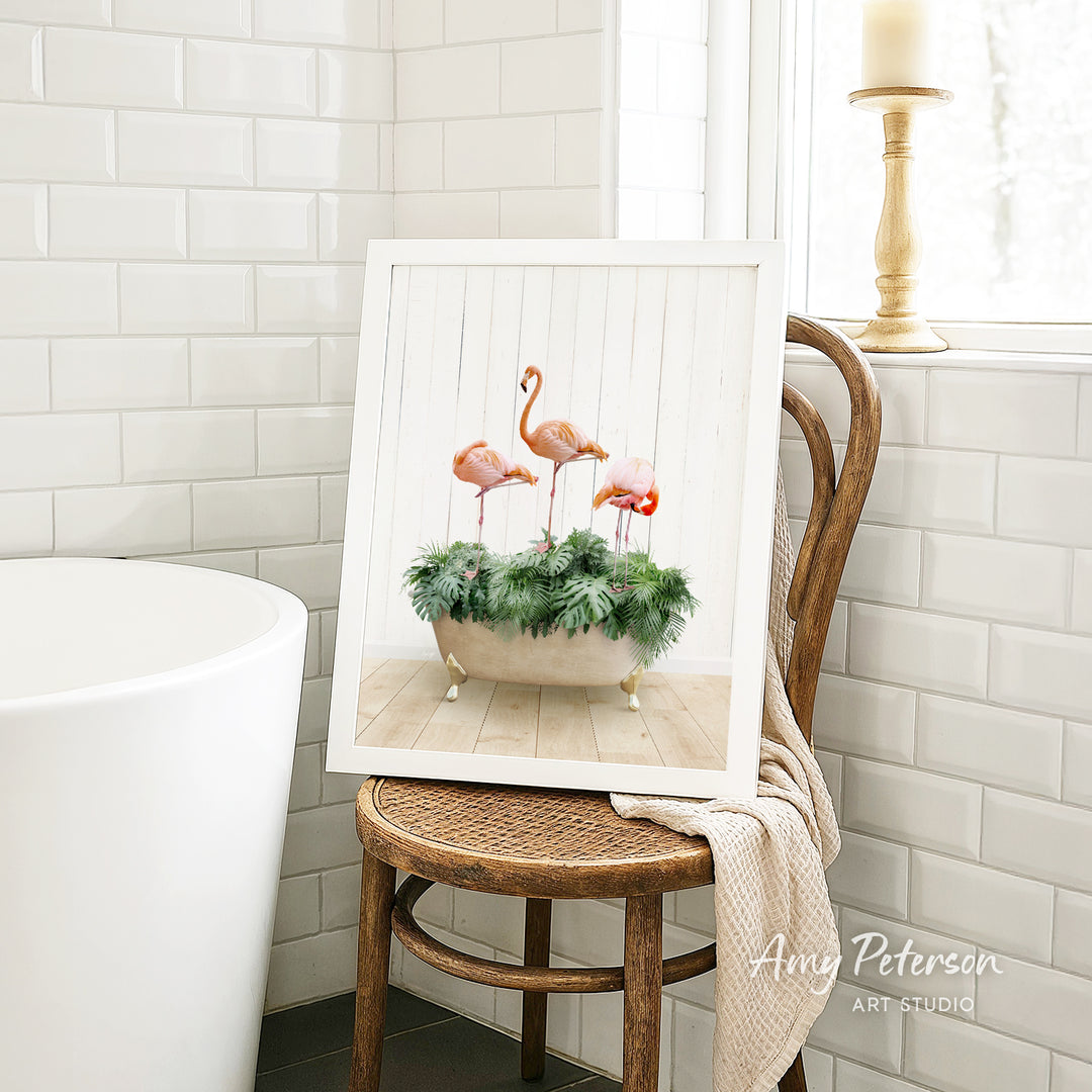 A framed image of three pink flamingos standing in a white vase filled with greenery, placed on a wooden chair in a bathroom setting.