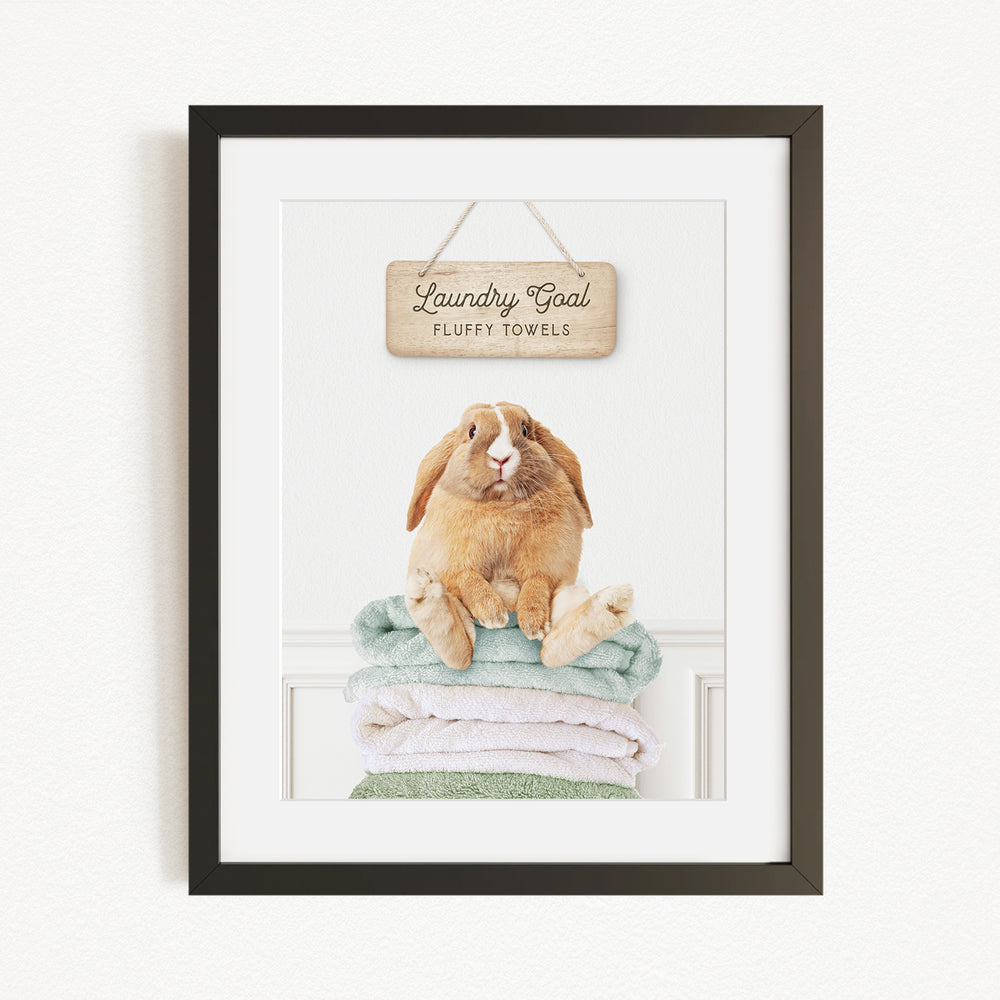 A framed image of a rabbit sitting on a stack of fluffy towels, with a wooden sign above that reads "Laundry goal Fluffy towels".
