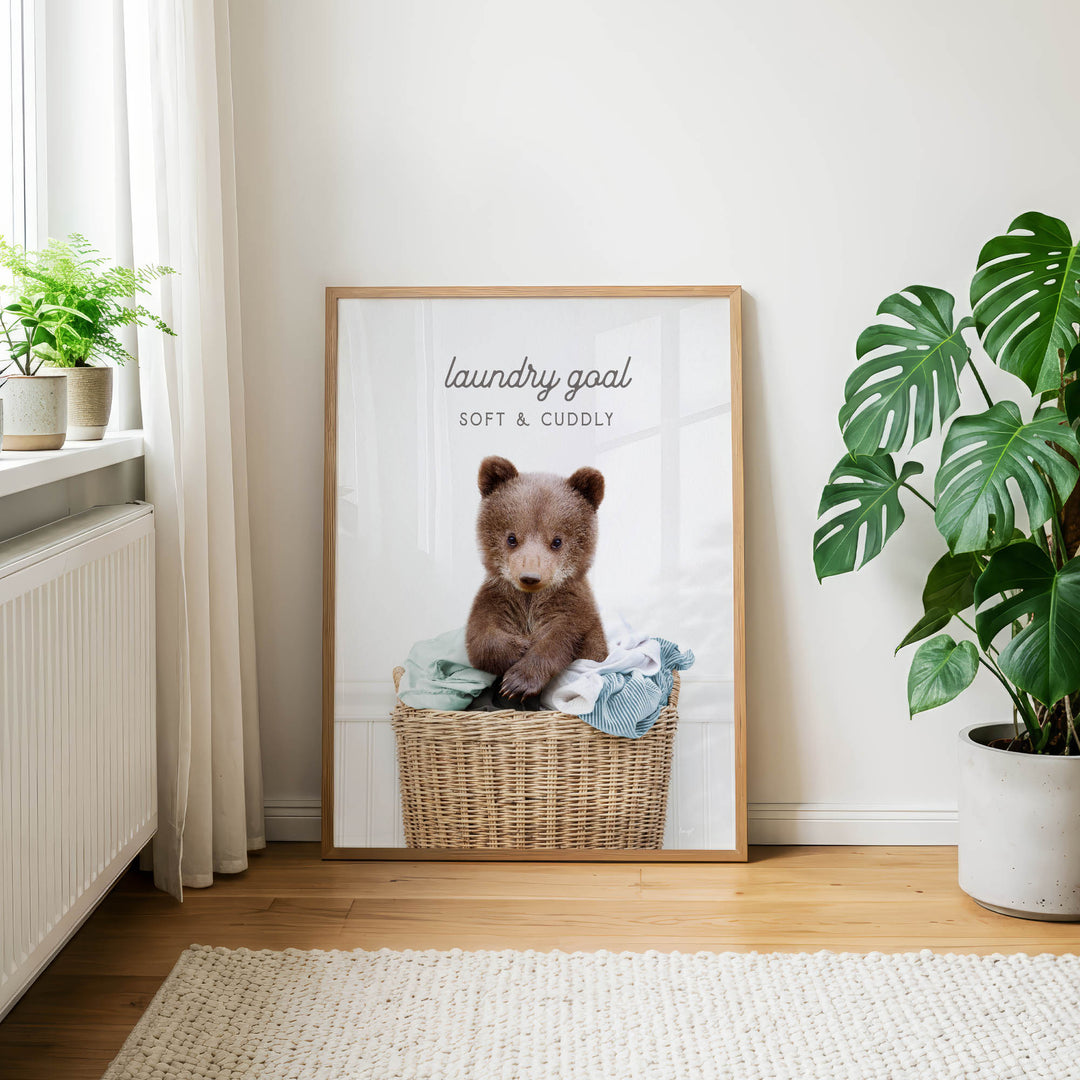 A framed poster of a brown bear sitting in a wicker basket, with the text "laundry goal soft & cuddly" on it, is displayed on a wall in a room with a window, plants, and a rug.