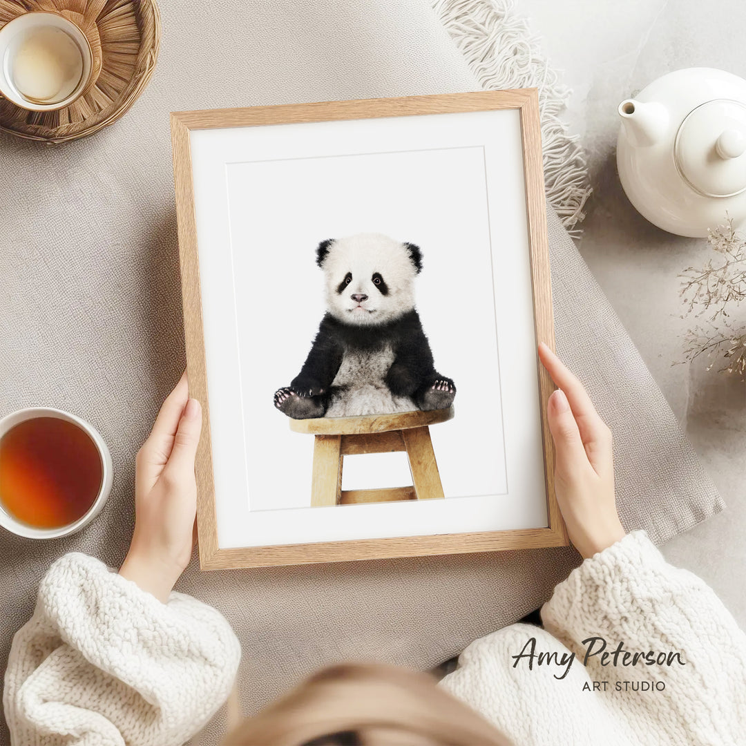 A person is holding a framed picture of a panda bear sitting on a wooden stool, with a teapot, a cup of tea, and a bowl of tea nearby.