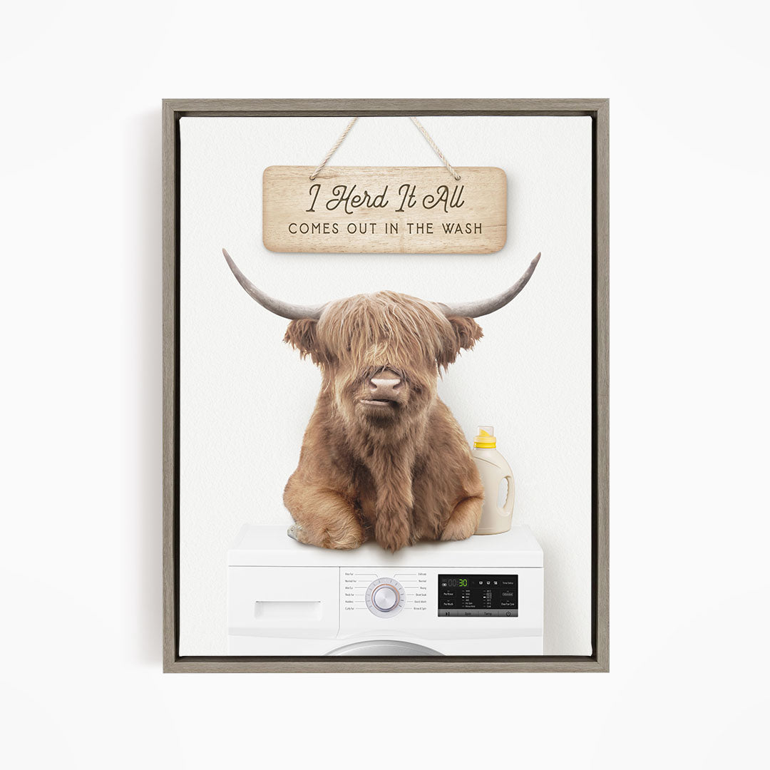 A brown cow with long horns sits on top of a white washing machine, with a wooden sign above it that says "I Herd It All Comes out in the Wash".