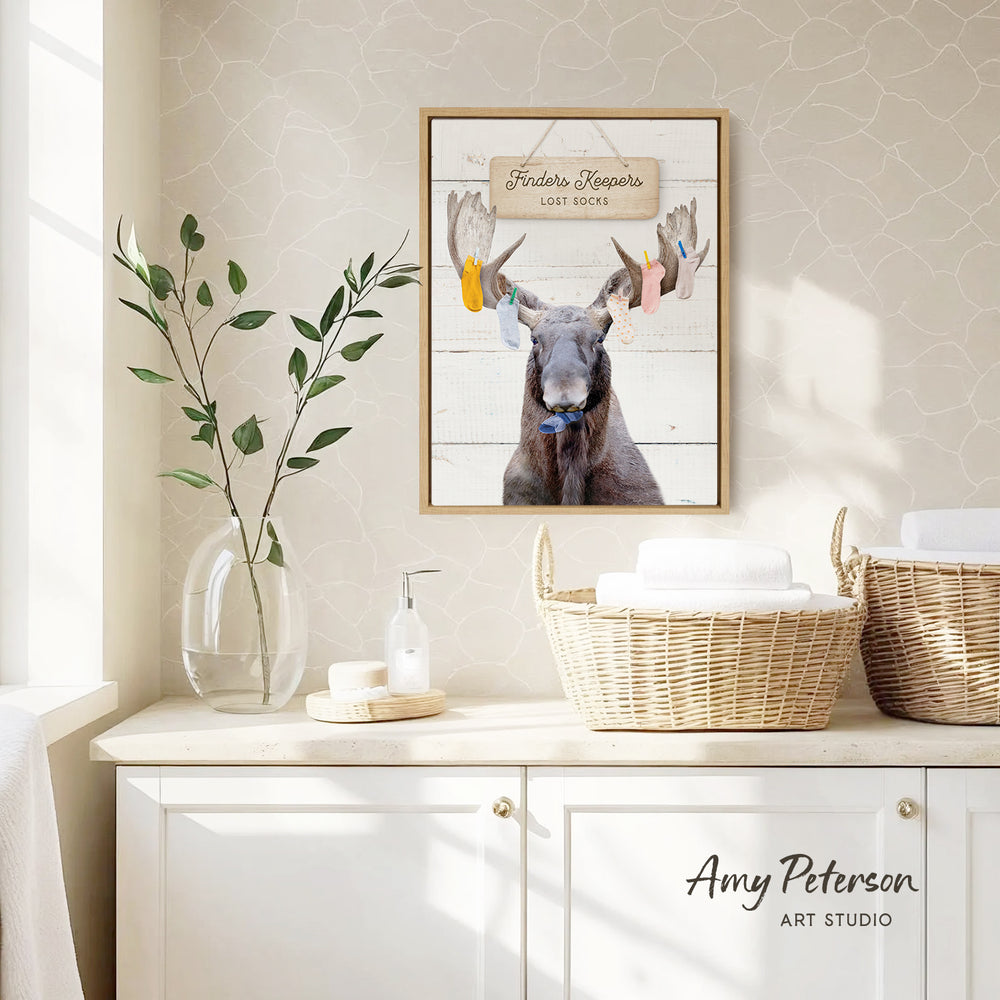 a framed picture of a moose with antlers, hanging on a wall above a white cabinet. The moose is wearing a blue bow tie. The cabinet has two woven baskets on top, and there is a vase with a plant on the counter. The wall has a patterned wallpaper.