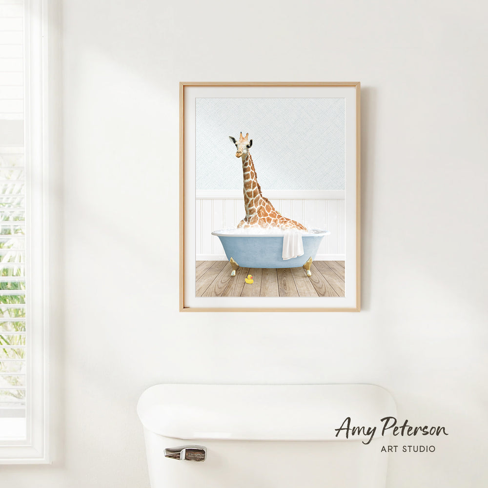 A framed illustration of a giraffe sitting in a bathtub, with a towel draped over its neck, is hanging on a white wall.