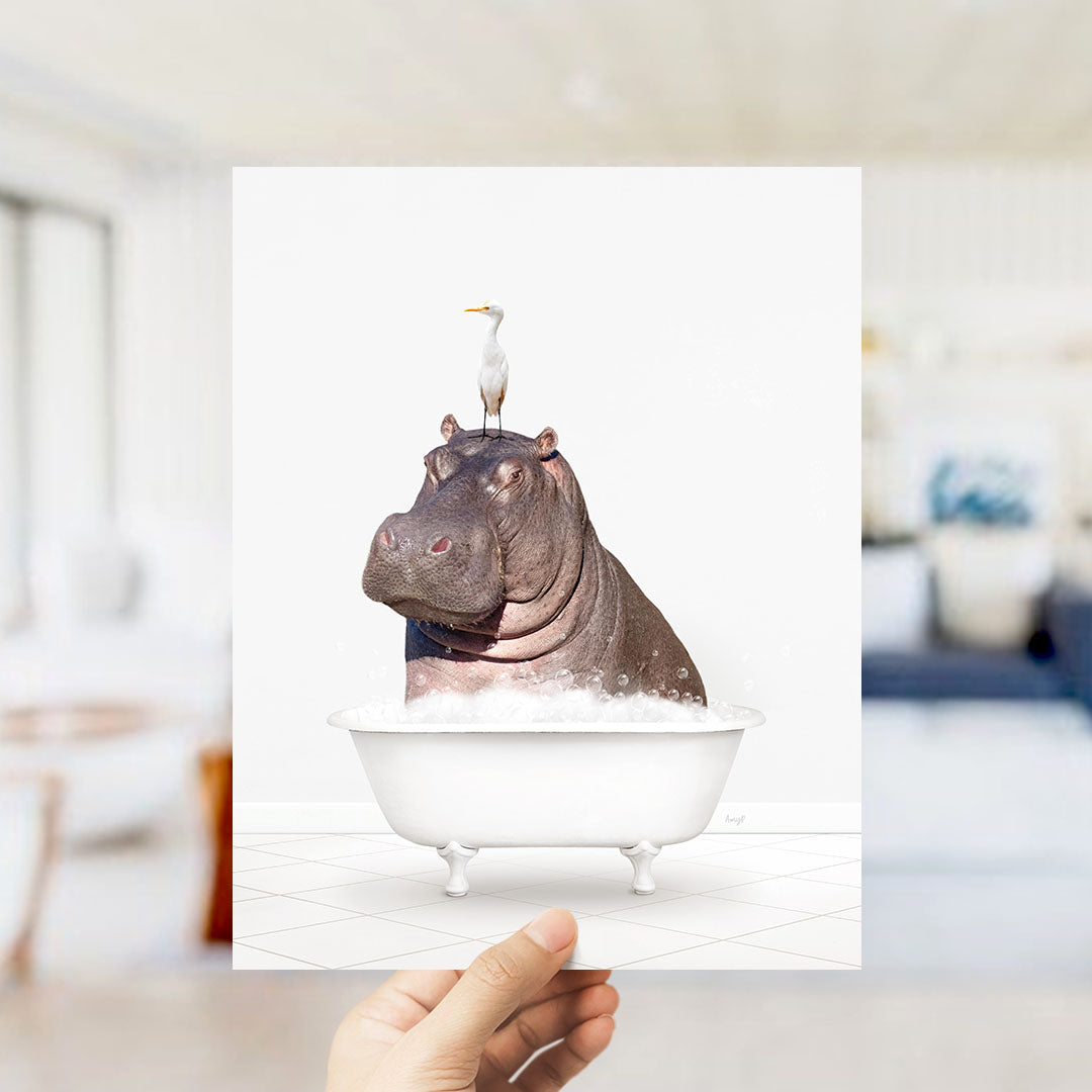 A hand holds up a framed image depicting a hippopotamus sitting in a bathtub, with a seagull perched on its head.