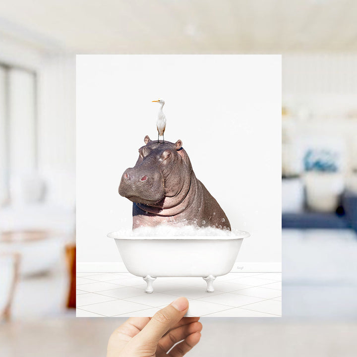 A hand holds up a framed image depicting a hippopotamus sitting in a bathtub, with a seagull perched on its head.