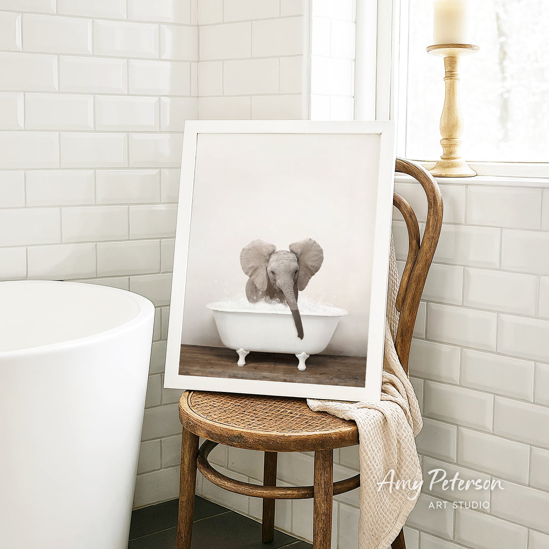 A framed photograph of a baby elephant sitting in a bathtub is displayed on a wooden chair in a bathroom setting.