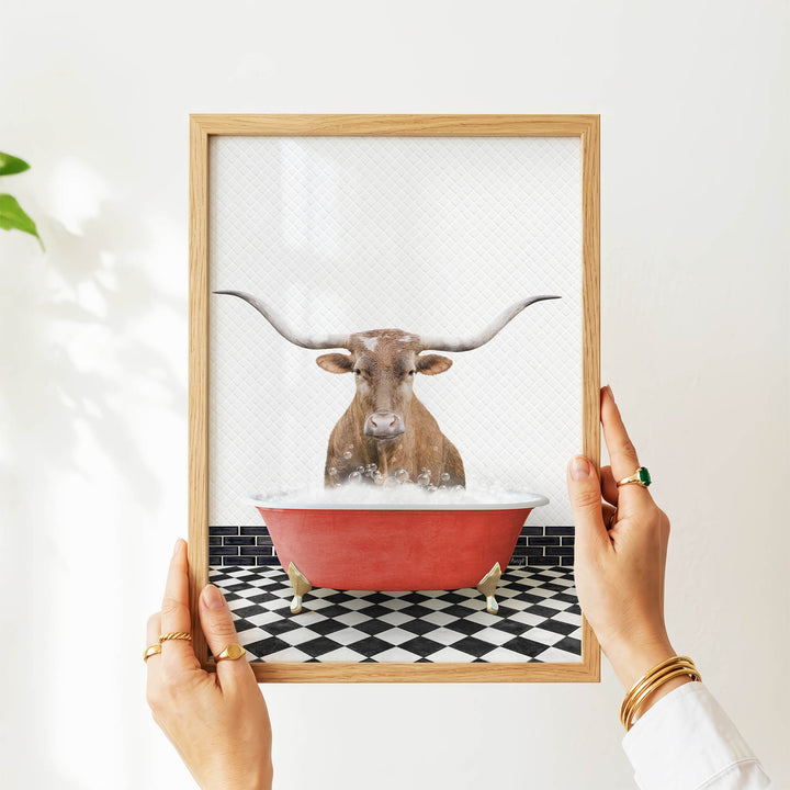 A person's hands are holding a framed picture of a bull with long horns, which is sitting in a red bathtub filled with water.