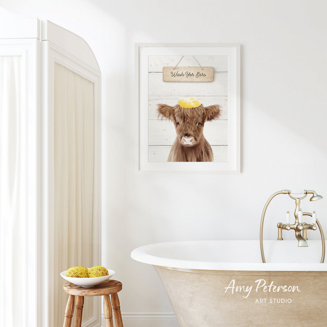 a bathroom with a framed picture of a cow on the wall, a wooden stool, a bowl of fruit, and a bathtub with a faucet.