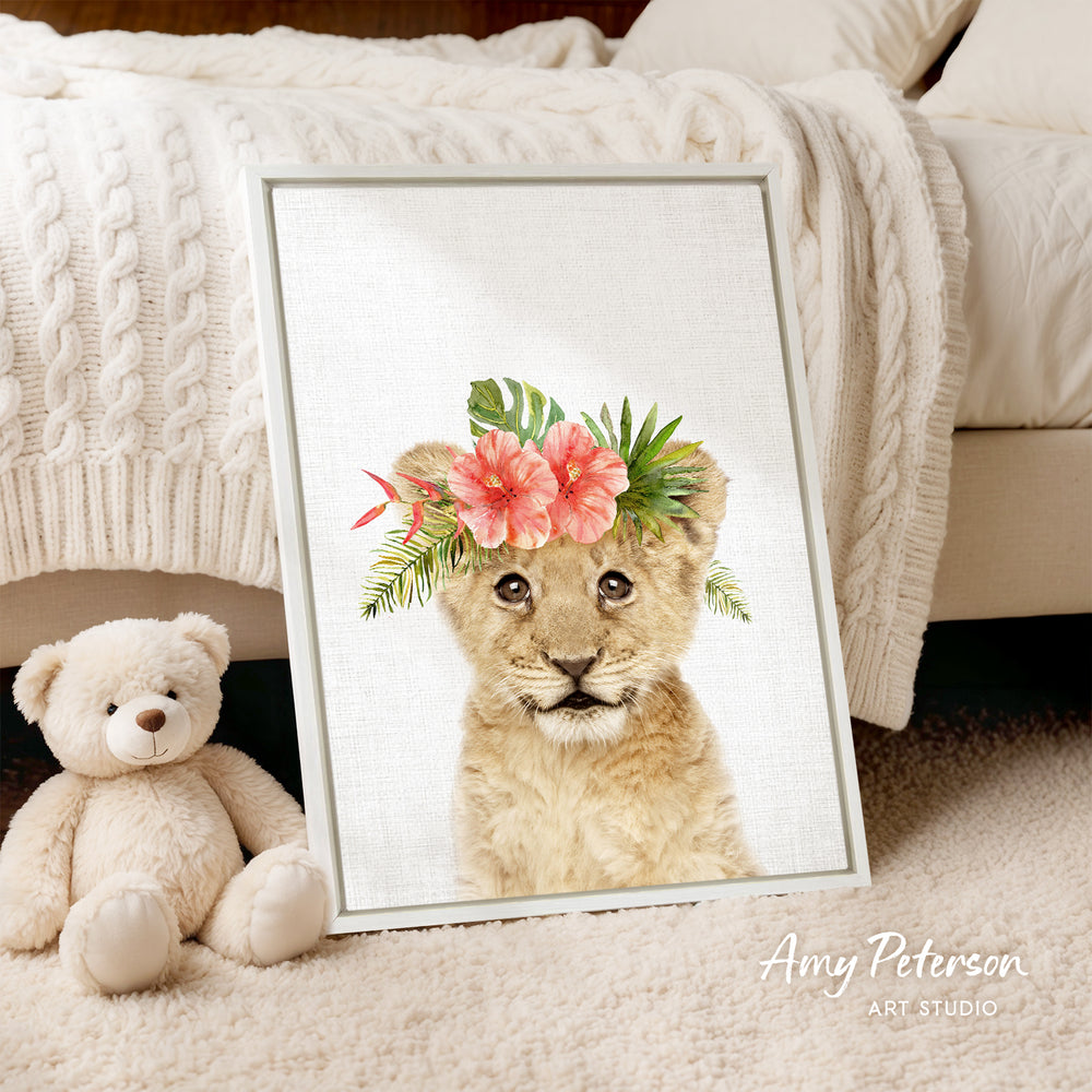 A framed print of a young lion wearing a floral crown is displayed on a white bedspread, accompanied by a teddy bear.