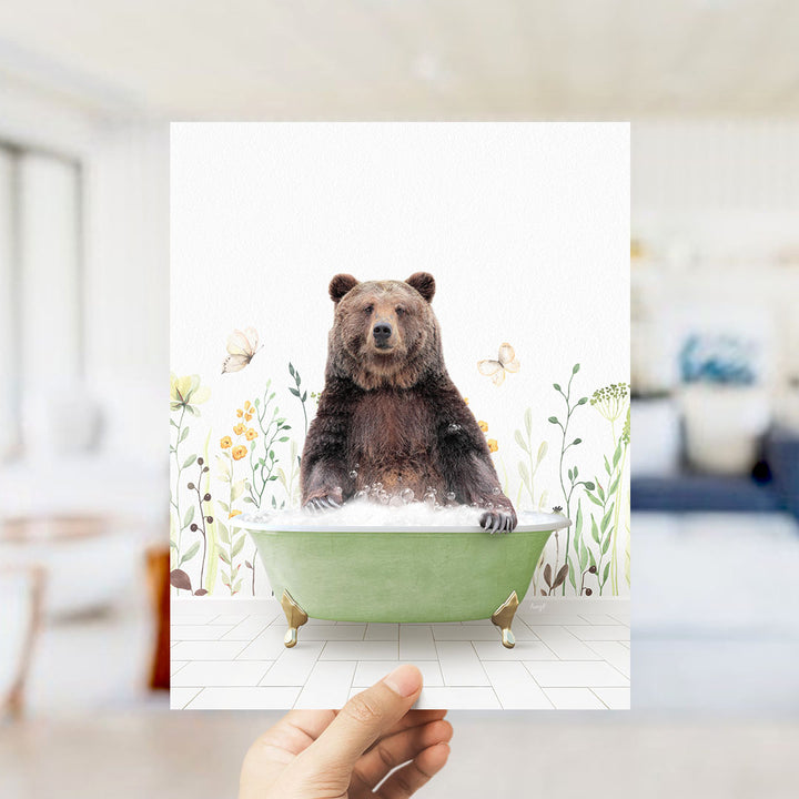 A hand holds up a framed image of a brown bear sitting in a green bathtub, surrounded by flowers and butterflies.