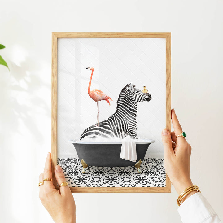 A person's hands are holding a framed picture of a zebra sitting in a bathtub, with a flamingo standing on its back.