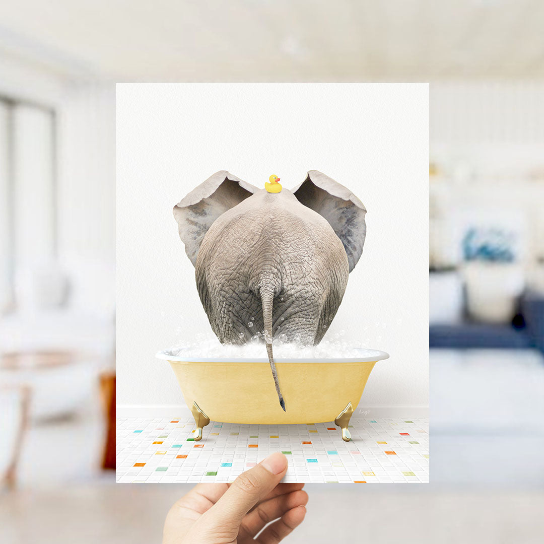 A hand holds up a framed image depicting an elephant sitting in a yellow bathtub, with a rubber duck perched on its head.