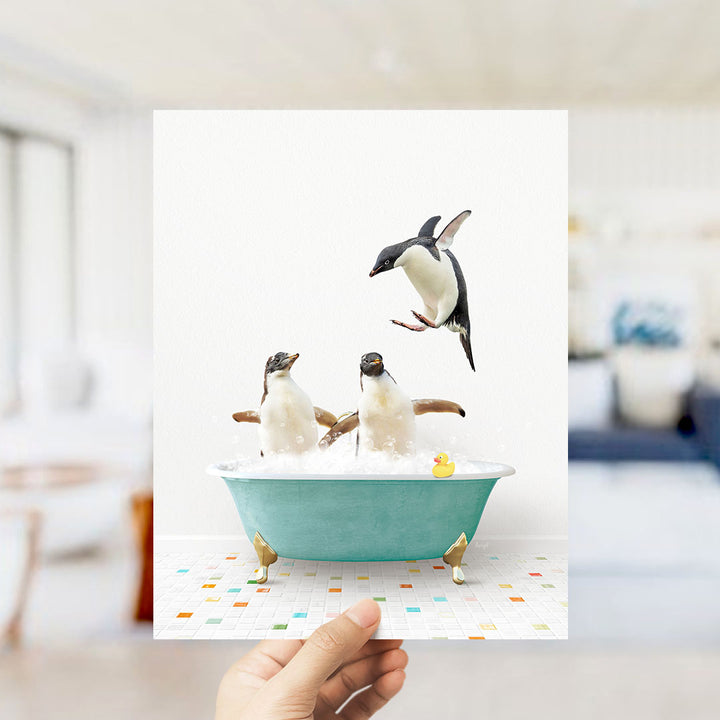 A hand holds up a framed image depicting two penguins sitting in a bathtub filled with bubbles, with one penguin flying towards the other.