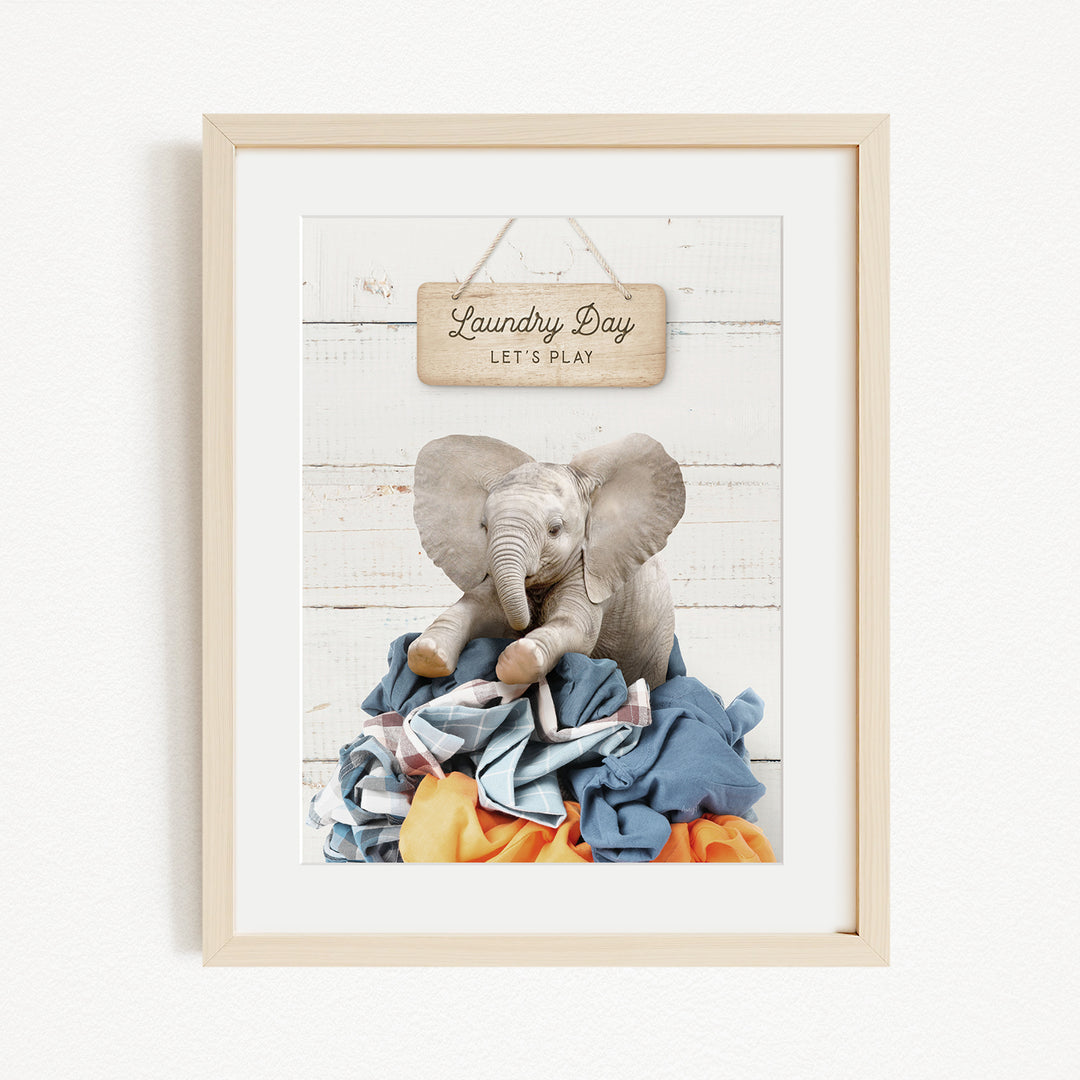 A framed image of a stuffed elephant sitting on a pile of clothes, with a wooden sign above it that says "Laundry Day Let's Play".