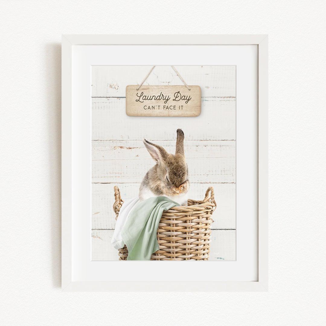 A framed image of a rabbit sitting in a wicker basket, with a wooden sign above it that says "Laundry Day Can't Face It".