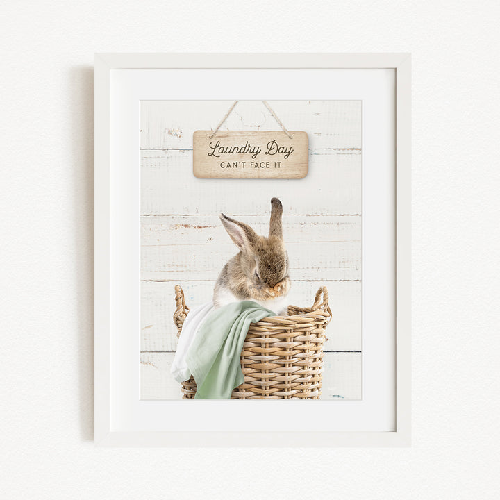 A framed image of a rabbit sitting in a wicker basket, with a wooden sign above it that says "Laundry Day Can't Face It".