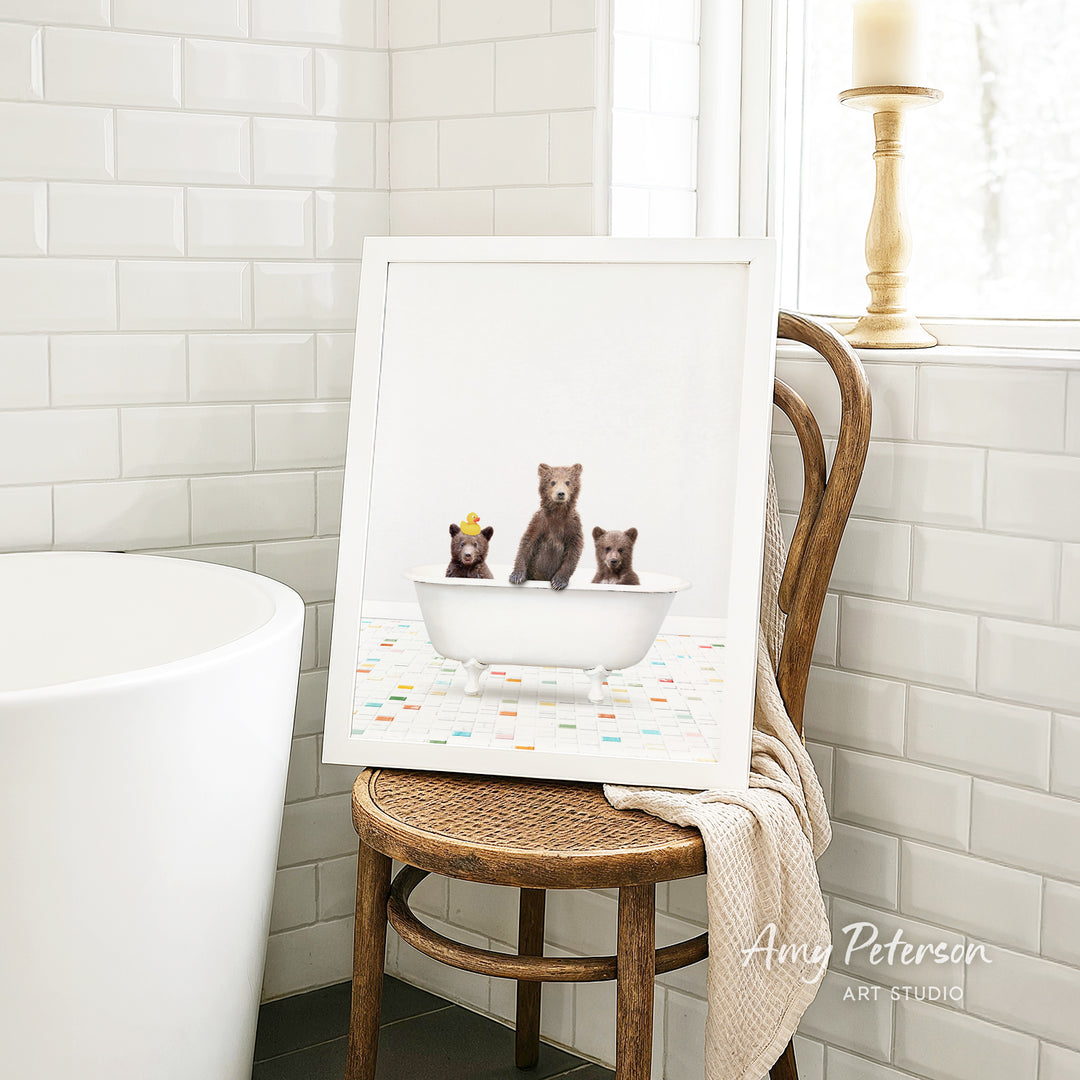 A framed image of three bears sitting in a bathtub, with a towel draped over the back of a chair in a bathroom setting.