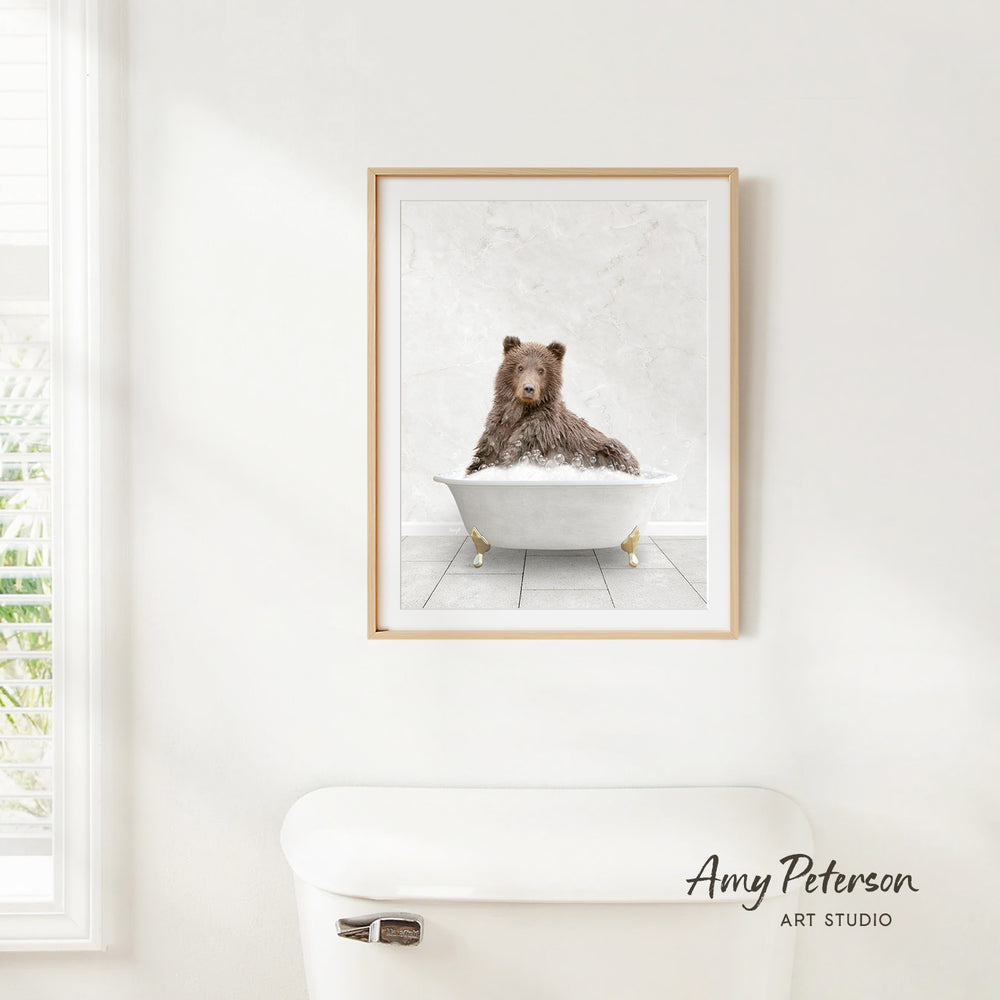 A framed image of a brown bear sitting in a white bathtub, with a window and a toilet visible in the background.