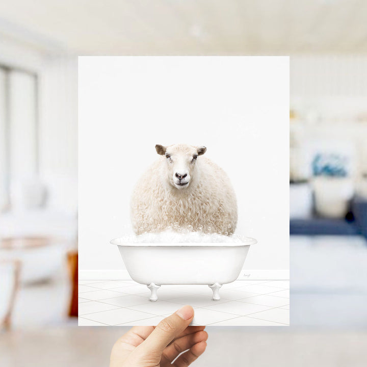 A hand holds up a framed image of a fluffy white sheep sitting in a white bathtub filled with bubbles.