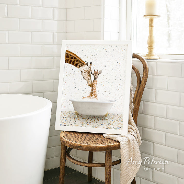A framed image of two giraffes standing in a bathtub, with one giraffe appearing to be bathing the other giraffe. The image is displayed on a wooden chair in a bathroom setting.