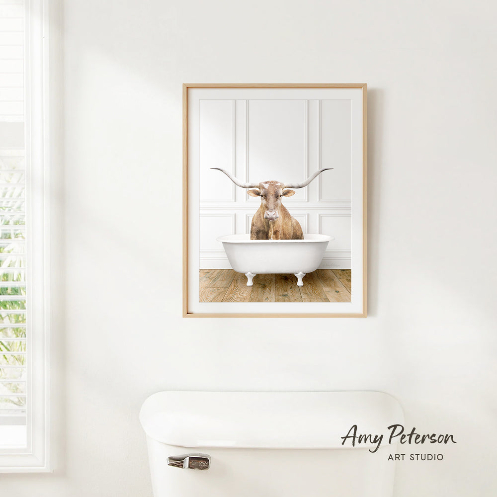 A framed image of a bull with long horns sitting in a white bathtub, hanging on a white wall.