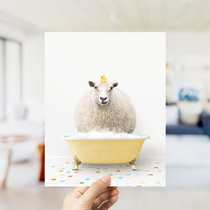 A hand holds up a framed image of a sheep sitting in a yellow bathtub, with a rubber duck on its head.