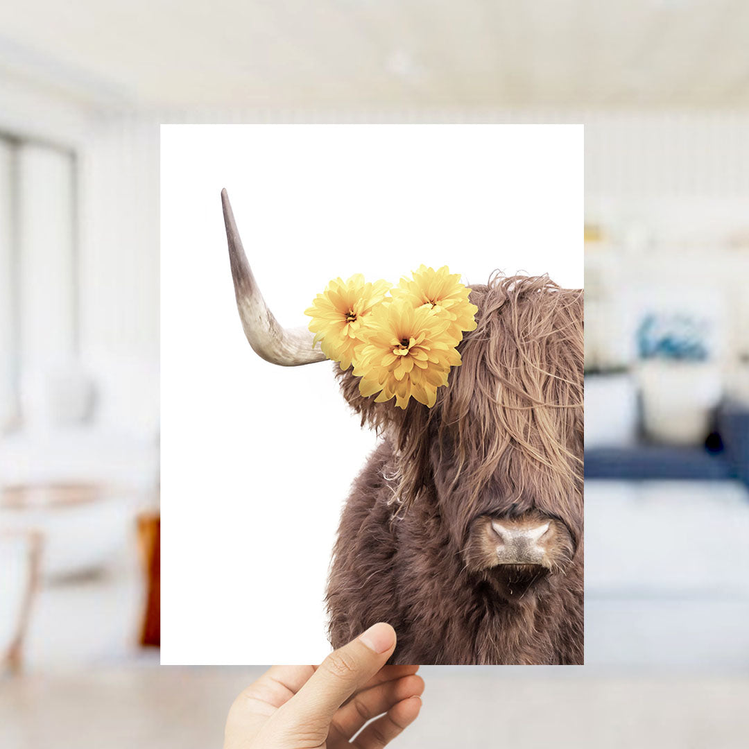 A hand holds up a framed image of a brown cow with yellow flowers on its horns.